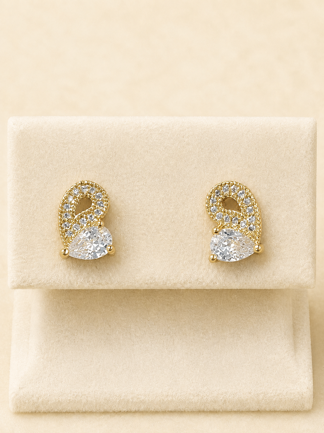 A pair of gold and crystal stud earrings displayed on a cream-colored jewelry stand against a warm beige background.
