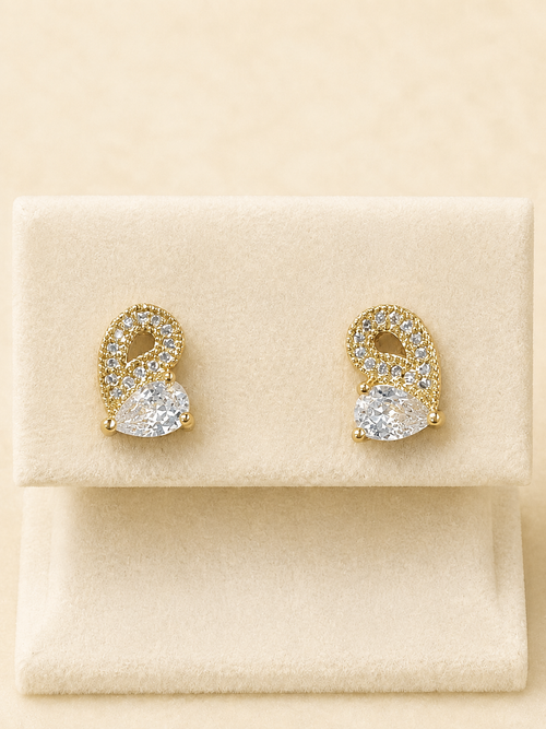 A pair of gold and crystal stud earrings displayed on a cream-colored jewelry stand against a warm beige background.