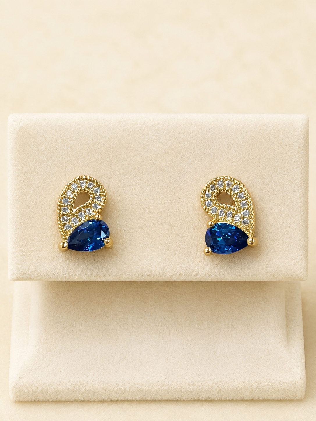 A pair of gold-tone stud earrings with pear-cut blue gemstones and pavé-set clear stones, displayed on a cream-colored jewelry box.