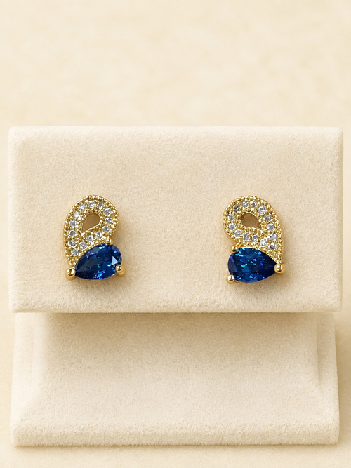 A pair of gold-tone stud earrings with pear-cut blue gemstones and pavé-set clear stones, displayed on a cream-colored jewelry box.