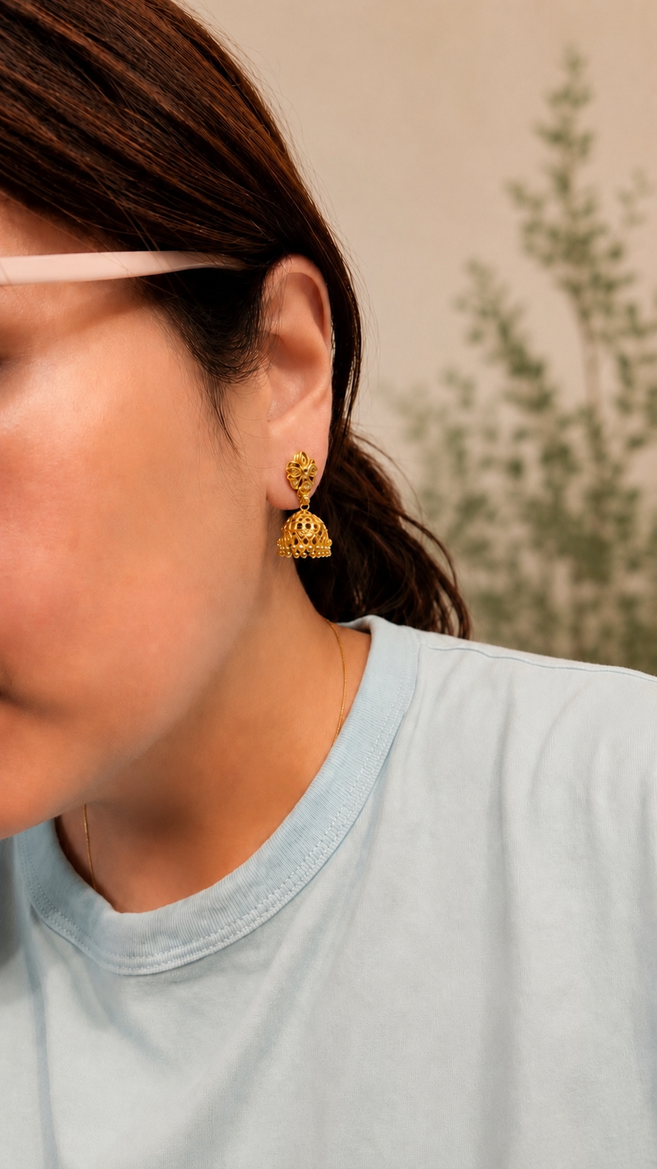 A close-up of a person wearing a small gold jhumka earring, with the side of their face, ear, and shoulder visible against a soft beige background with blurred greenery.