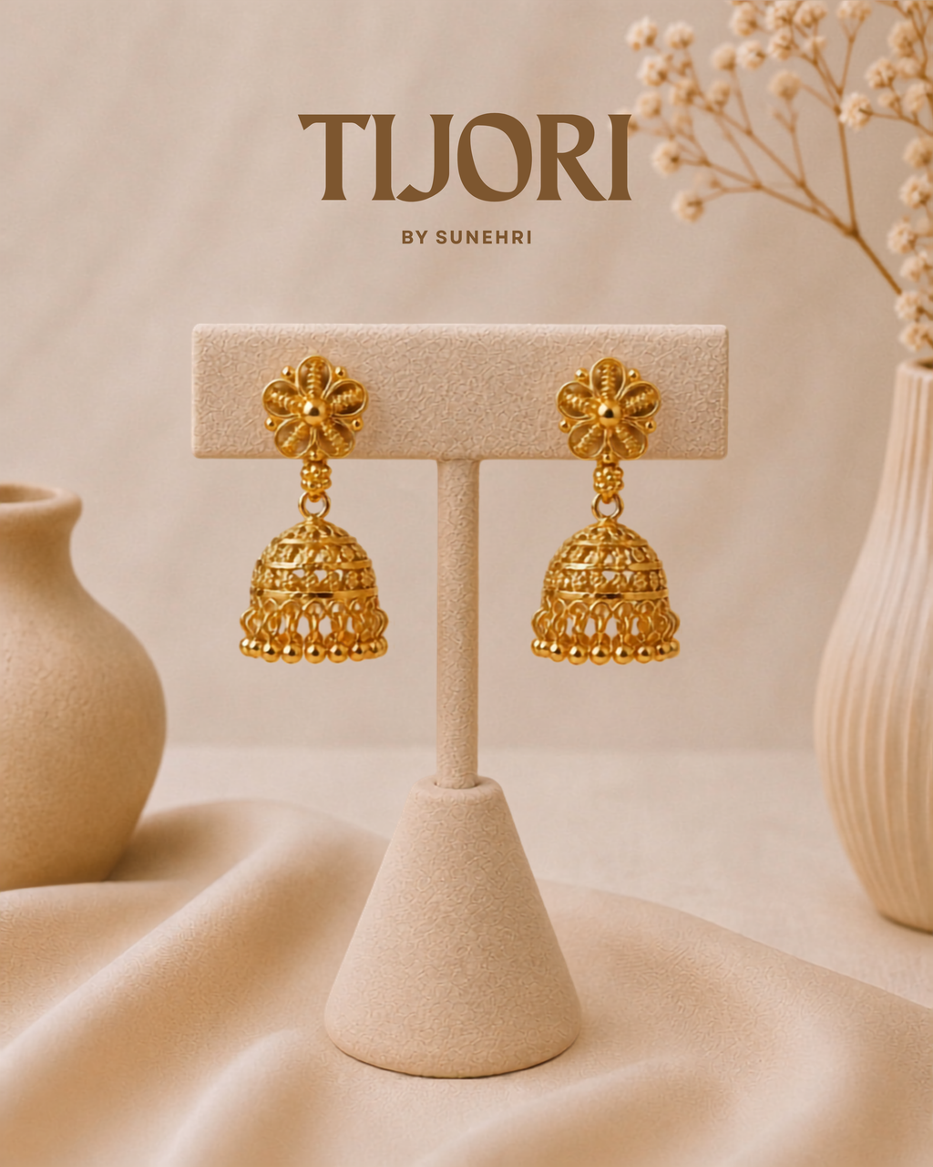 A pair of gold-toned traditional jhumka earrings displayed on a beige jewelry stand. The background is a soft beige fabric with decorative ceramic vases and dried flowers, and the brand name 'TIJORI BY SUNEHRI' is printed at the top.