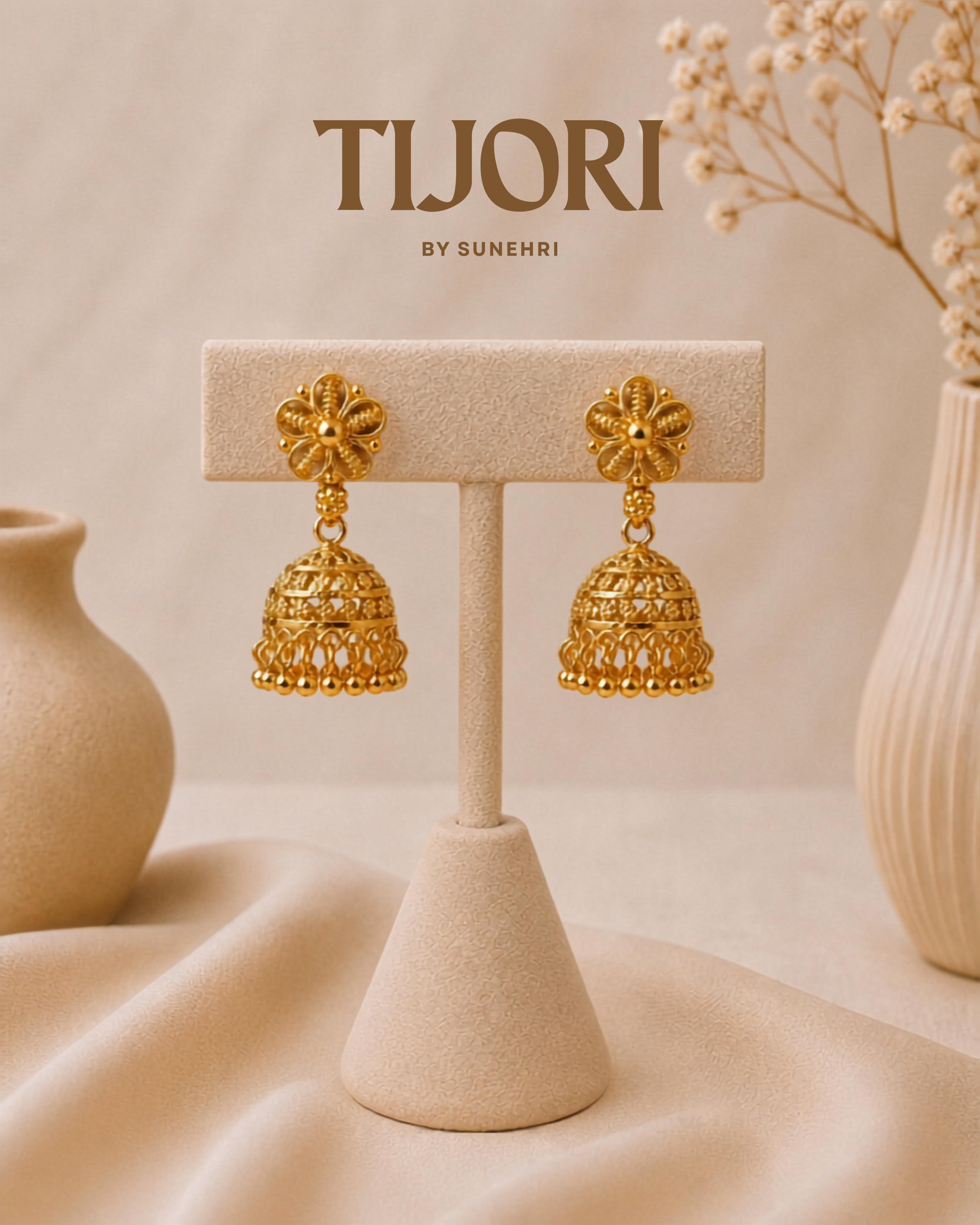 A pair of gold-toned traditional jhumka earrings displayed on a beige jewelry stand. The background is a soft beige fabric with decorative ceramic vases and dried flowers, and the brand name 'TIJORI BY SUNEHRI' is printed at the top.