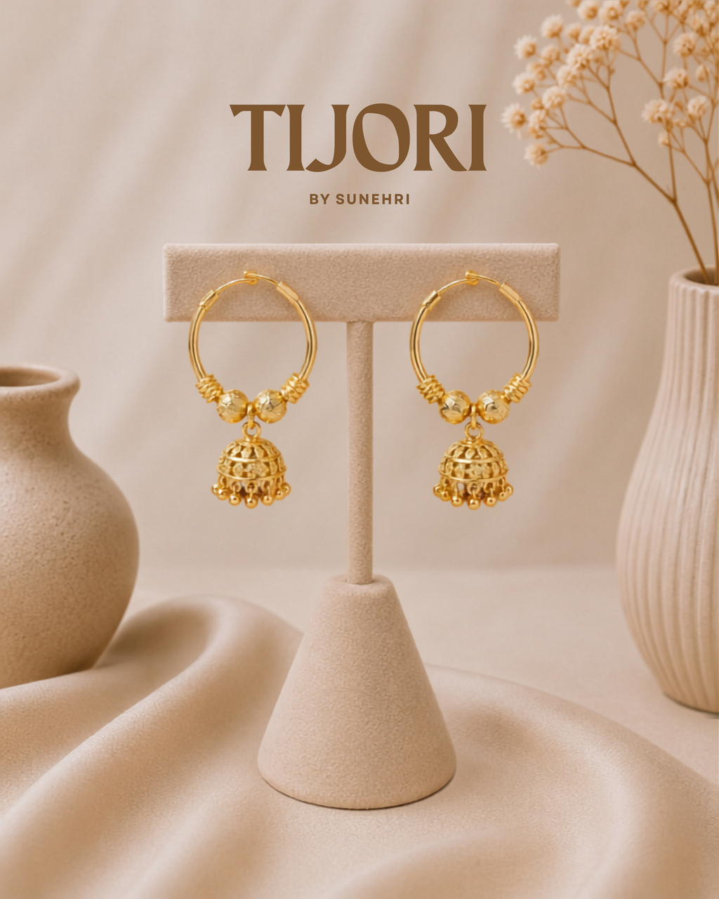 A pair of gold-toned hoop earrings with dangling bell-shaped charms and small bead details, displayed on a beige jewelry stand. The background is a soft beige studio setting with draped fabric, decorative ceramic vases, and dried flowers.
