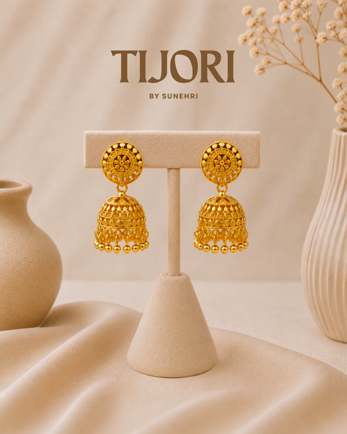 A pair of ornate gold-toned Jhumka earrings displayed on a beige jewelry stand. The background is a soft beige fabric with decorative ceramic vases and dried flowers, and the brand name 'TIJORI BY SUNEHRI' is printed at the top.
