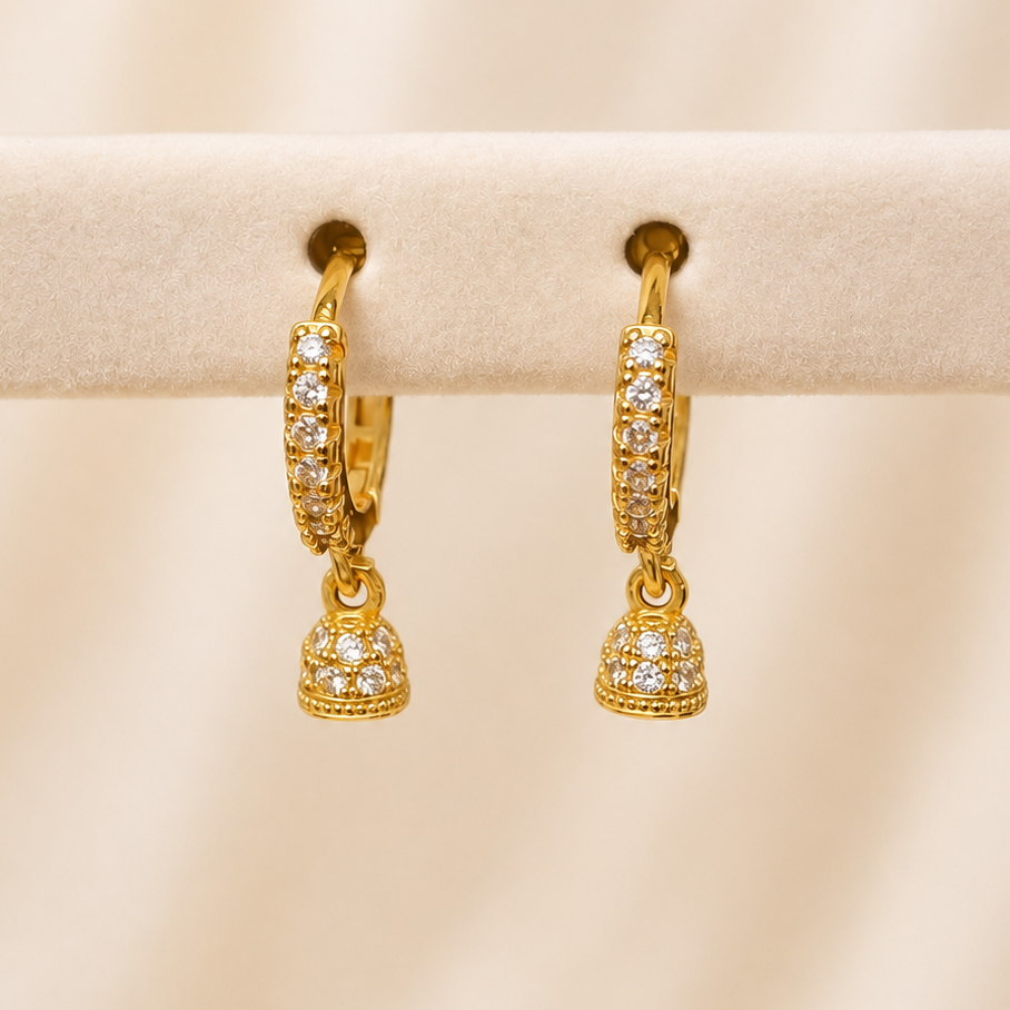 A pair of gold huggie hoop earrings adorned with pavé-set clear stones and a dangling bell-shaped charm, displayed on a cream-colored jewelry stand against a softly draped beige background.